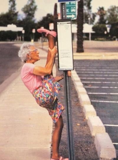 An older woman in colorful clothes stretching her leg on a bus stop sign
