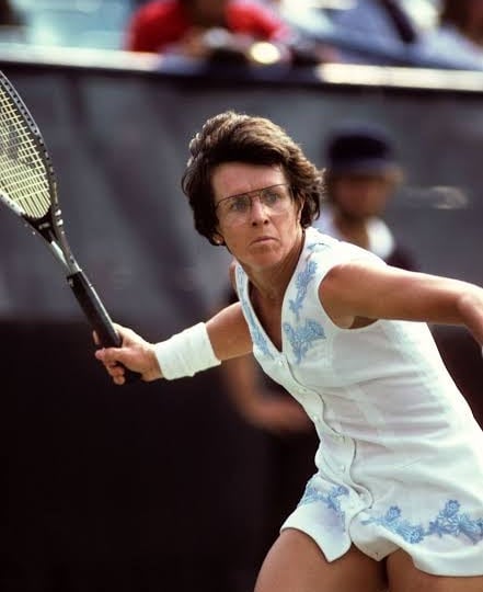 Billie Jean King in a white embroidered tennis dress, eyes on the ball, racquet back