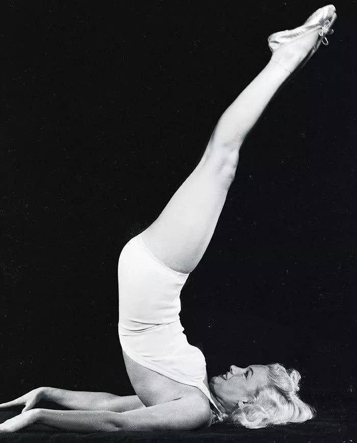 A woman in a white leotard in a shoulder-stand with her legs raised, in black and white